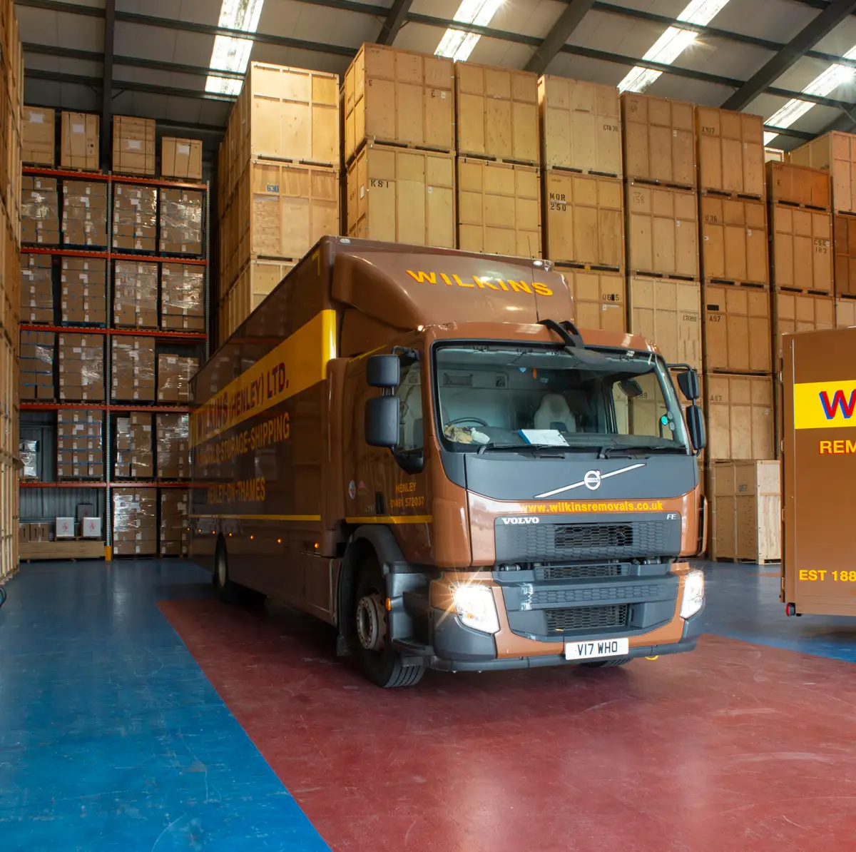 Large brown Wilkins Removals lorry with company branding and contact details is parked inside a warehouse. The grey warehouse features orange accents, whilst the scene highlights the reliable removals service.